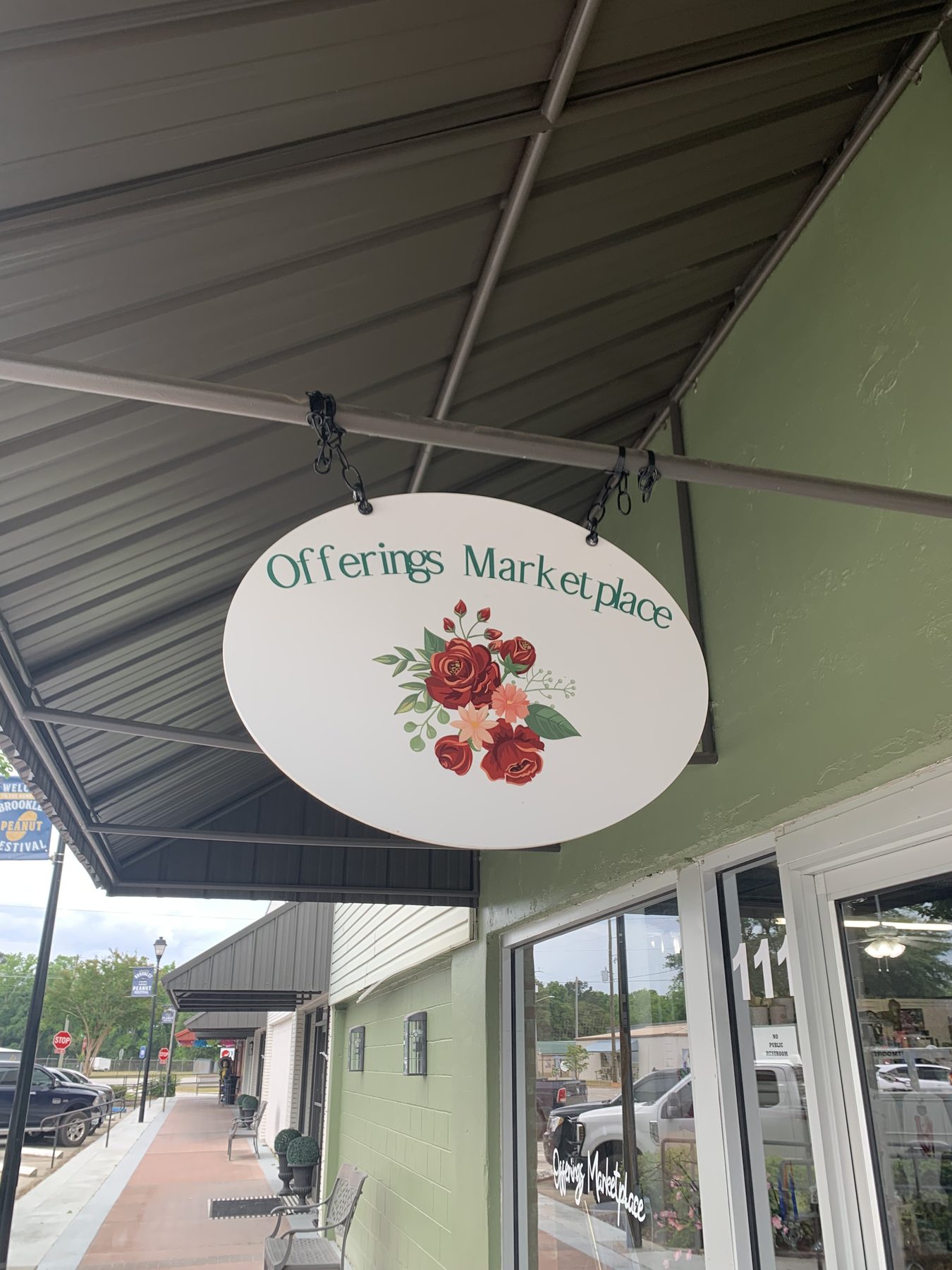 The Offerings Marketplace oval sign, painted with a rose bouquet, hanging beneath the storefront awning